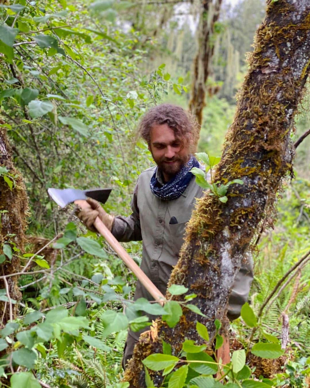 Man with axe in a lush forest clearing near mossy trees.