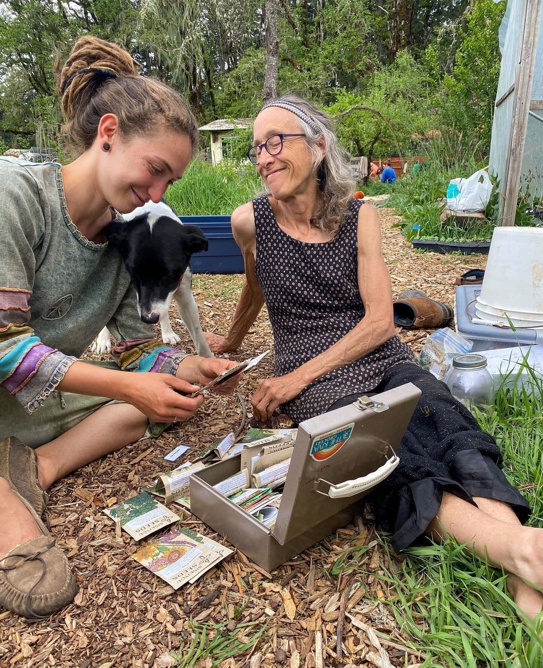 Two women with a dog exchanging cards and items outdoors at an eco village.