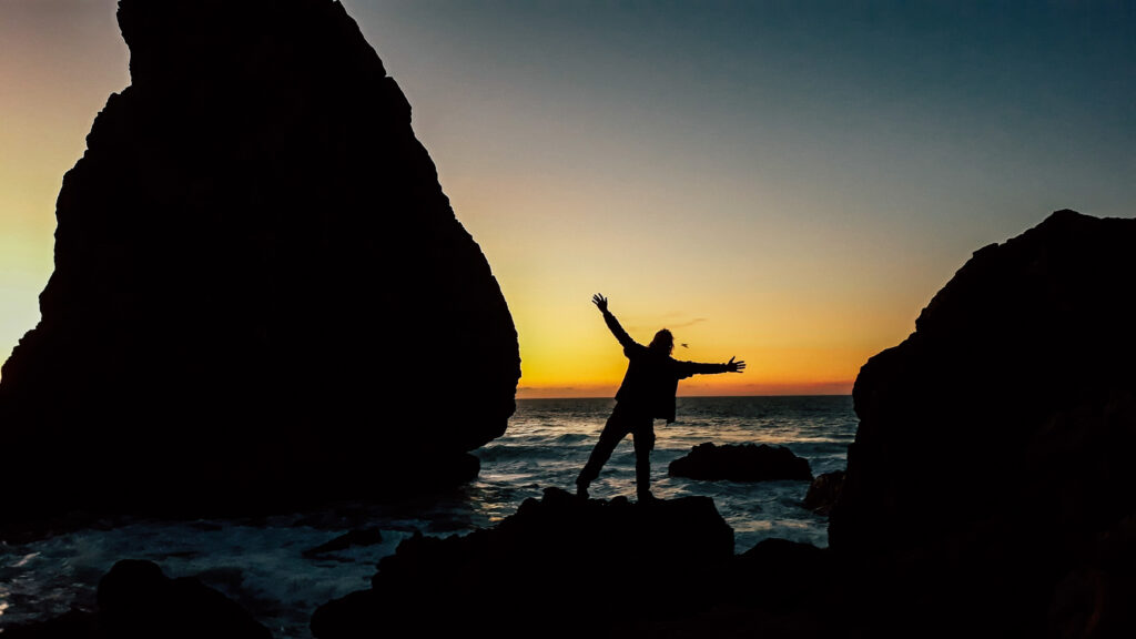 Person on rocky coastline at sunset with arms raised near sea.