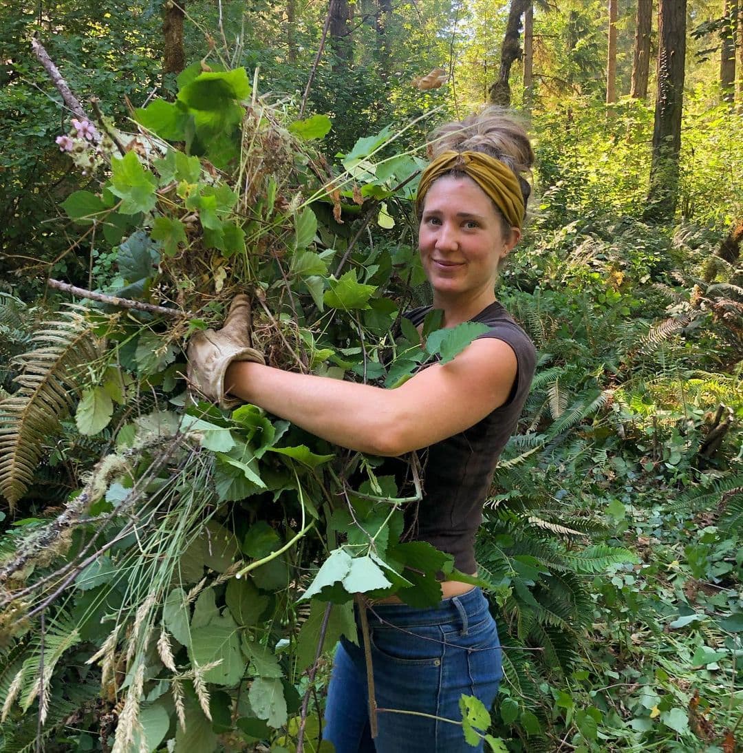 Woman in a forest holding a bundle of leaves and plants.