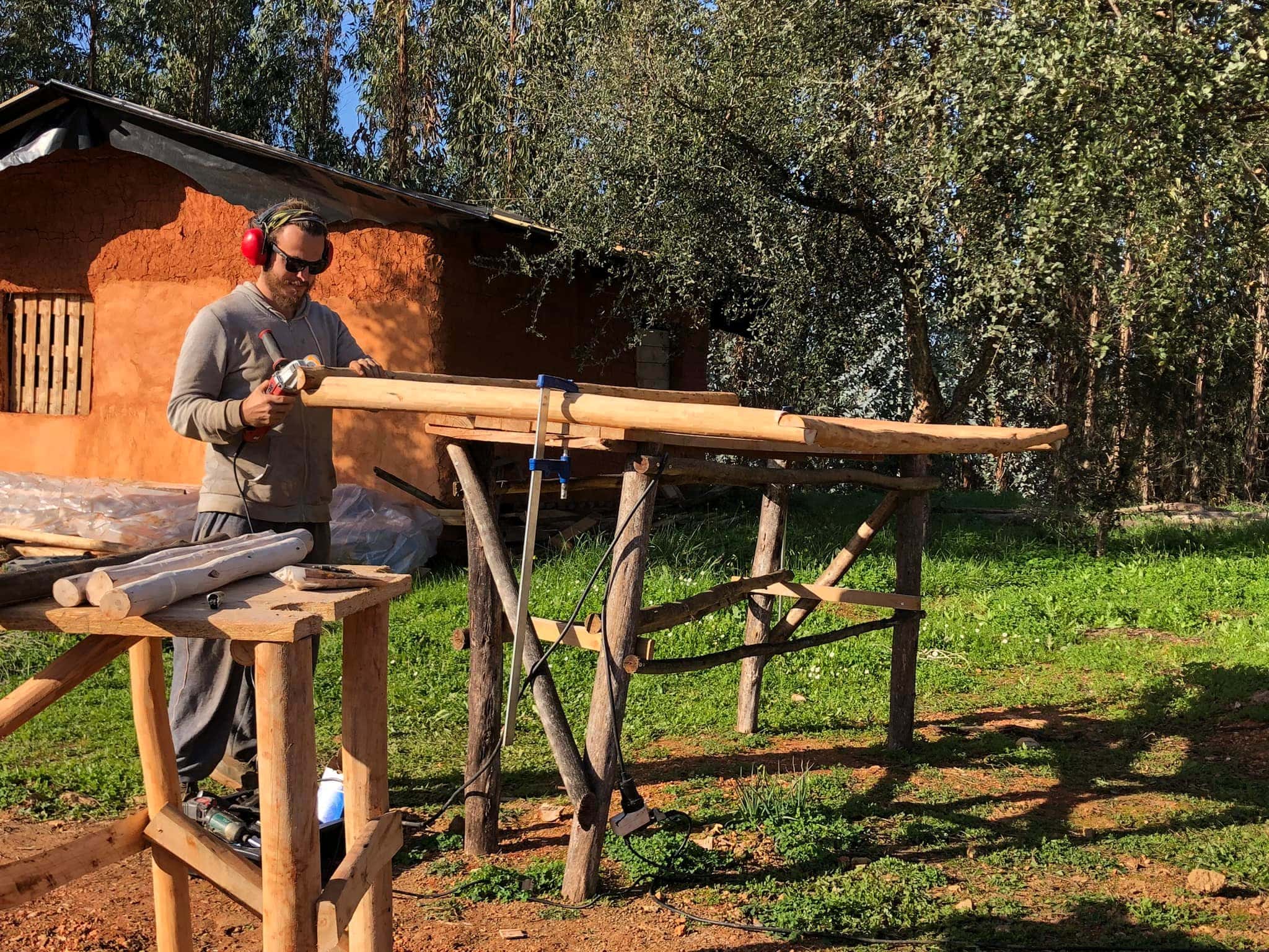 Person shaping a wooden beam outdoors at Monte do Aniche workshop with rustic tools.