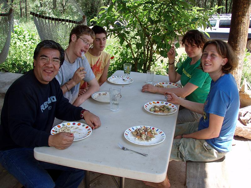 Group of six people sitting at a long table eating outdoors.