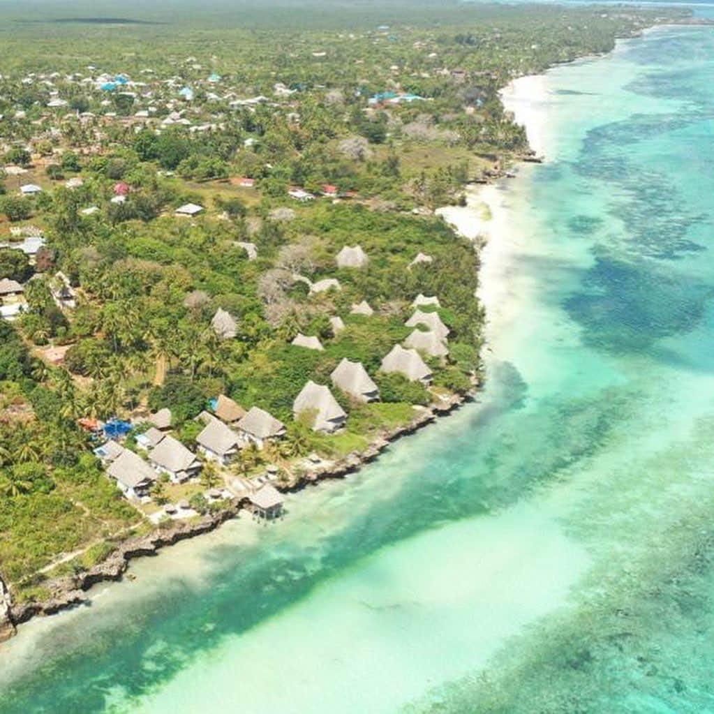 Aerial view of a tropical eco-village along a turquoise shoreline with thatched-roof huts and lush/W.