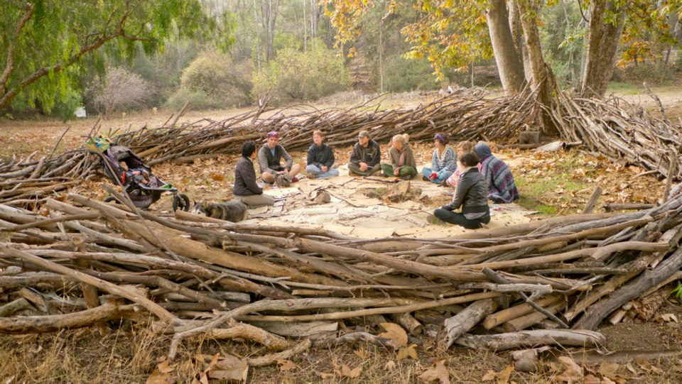 Global ecovillage network gathering around a communal fire circle in a forest.