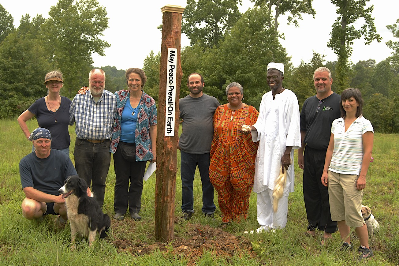 Group of diverse community members beside a wooden post with NG title in a grassy field, smiling.