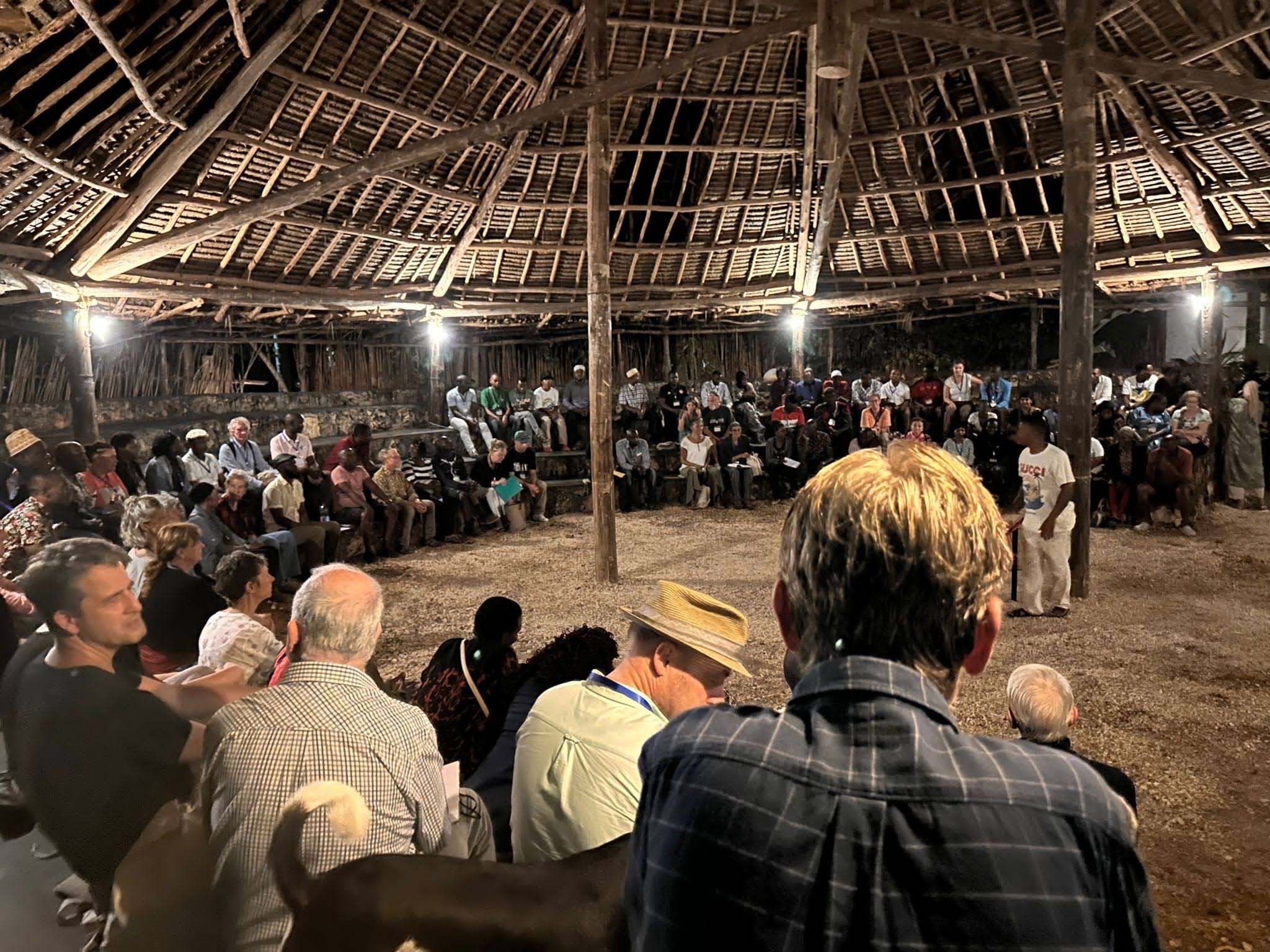 People gather in a circular open-air hut with a thatched roof and wooden posts at dusk.
