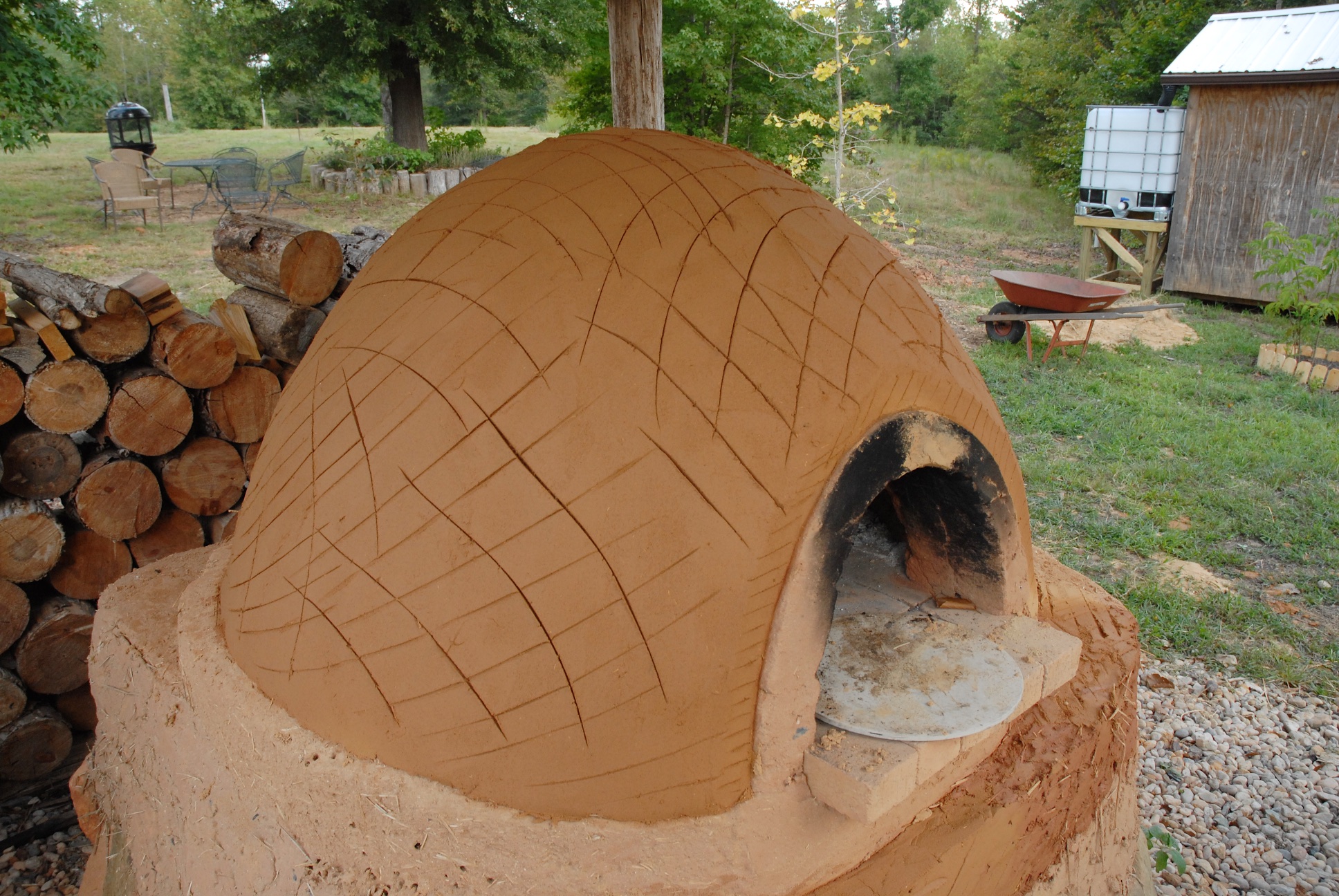 Clay wood-fired dome oven at ecovillage site with log pile and shed in background.