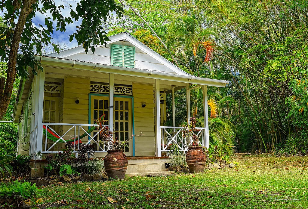 Yellow Caribbean cottage with a covered porch surrounded by lush tropical greenery.