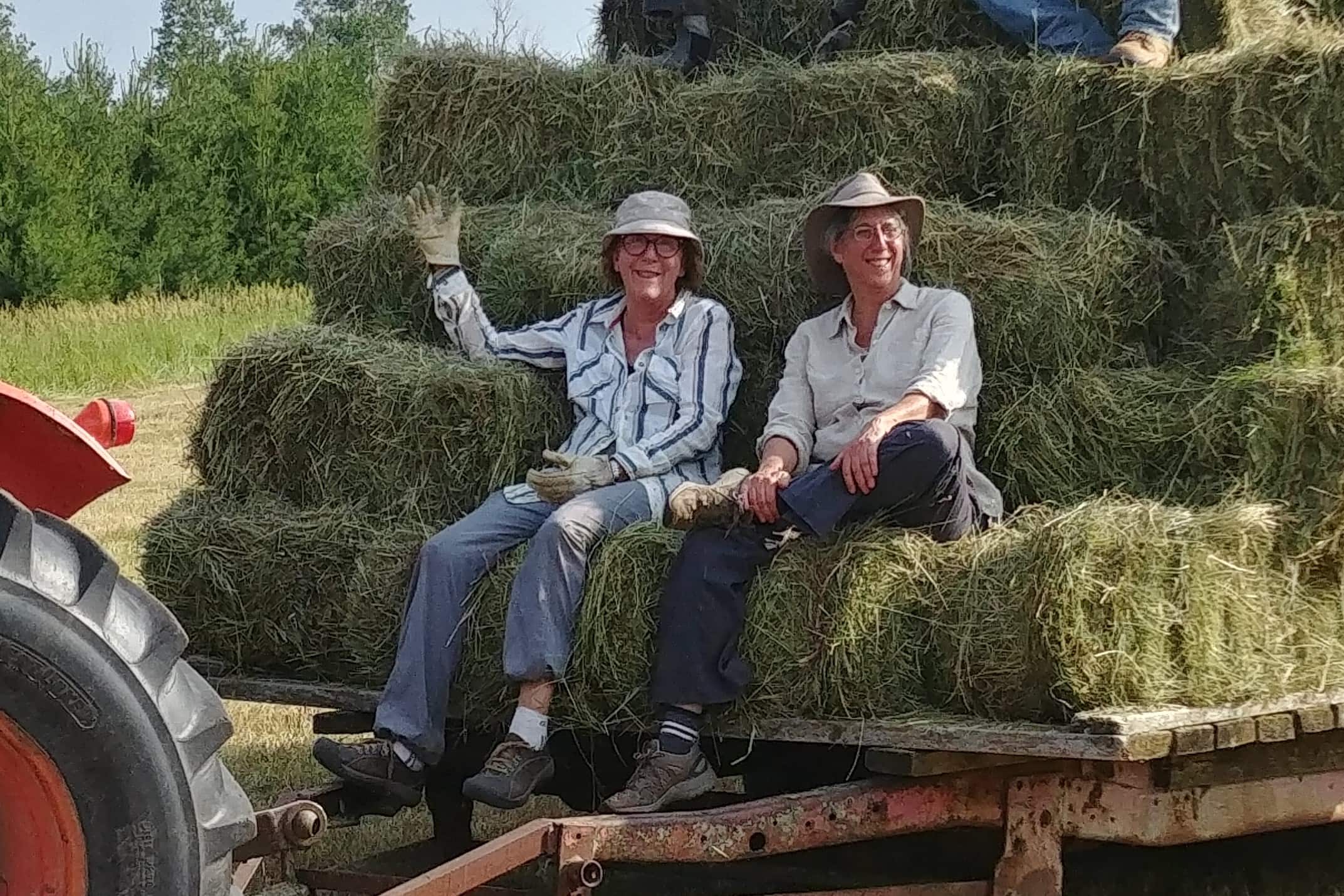 Two people sitting on a hay wagon with stacked hay bales in a field.