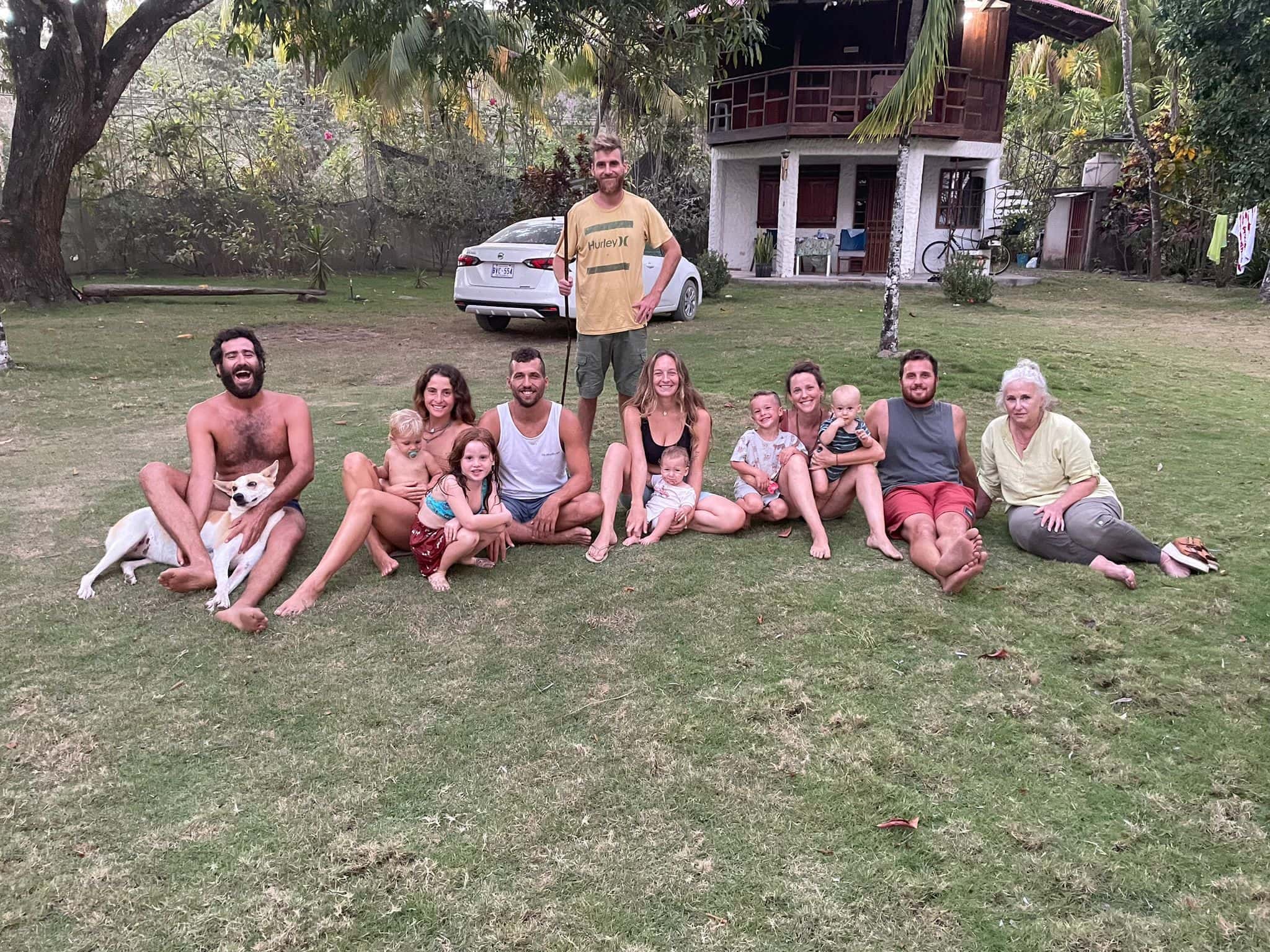 Group of diverse people sitting on grass in front of a house at Beija Flor Village, outdoor setting.