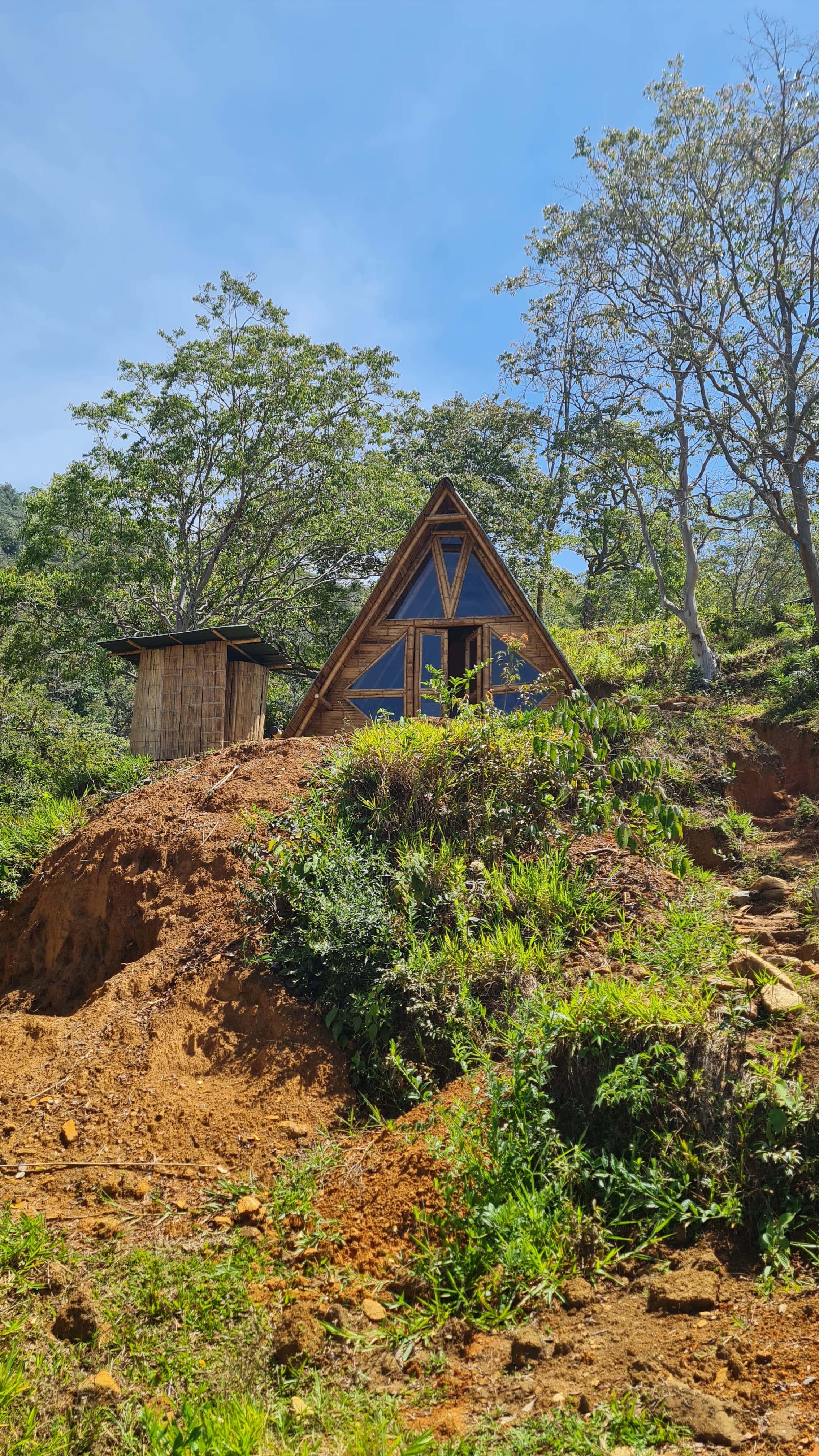 A-frame eco cabin perched on a dirt hillside with surrounding trees under a blue sky.
