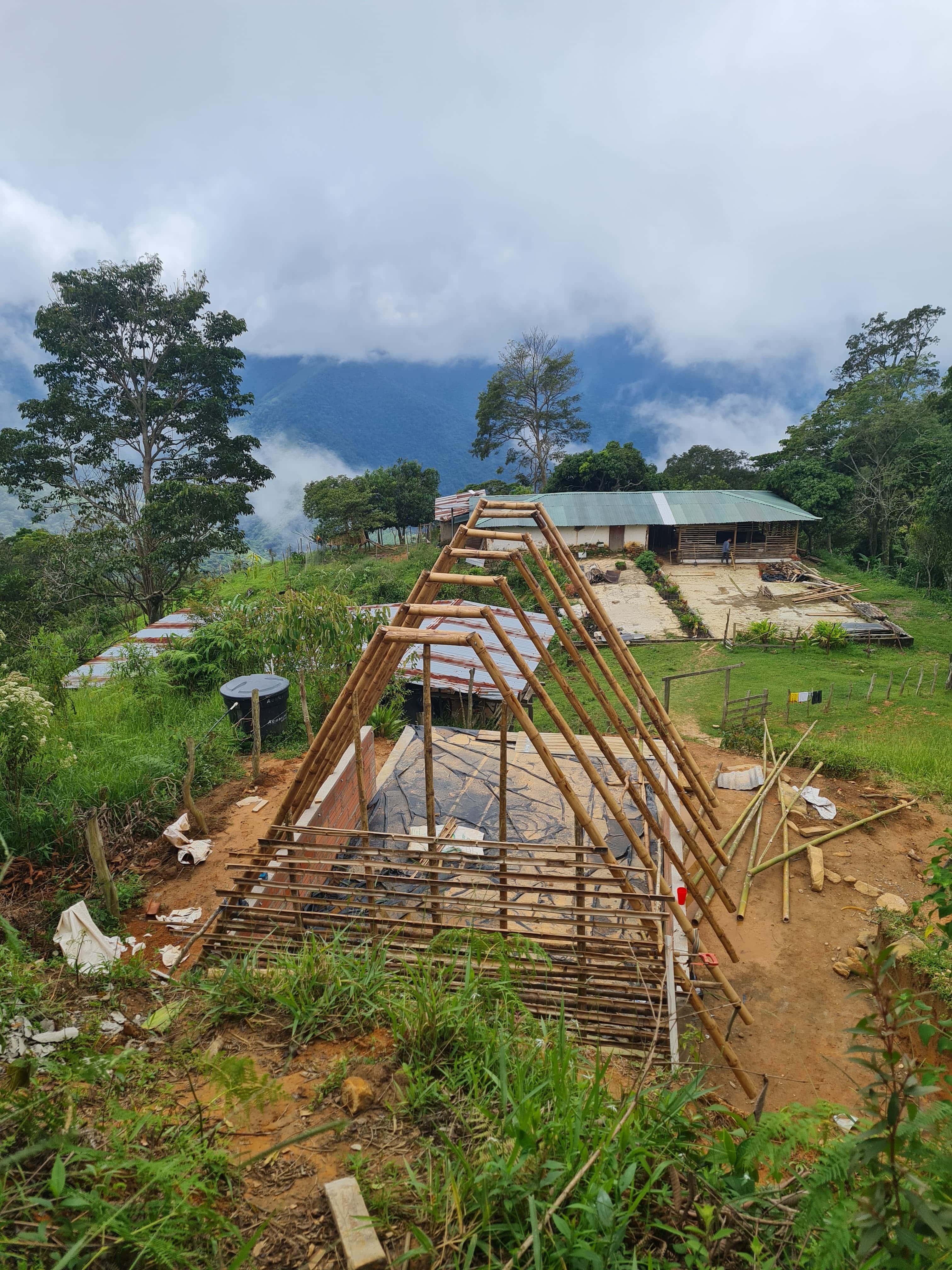 Timber triangular frame under construction with hills and huts in background.