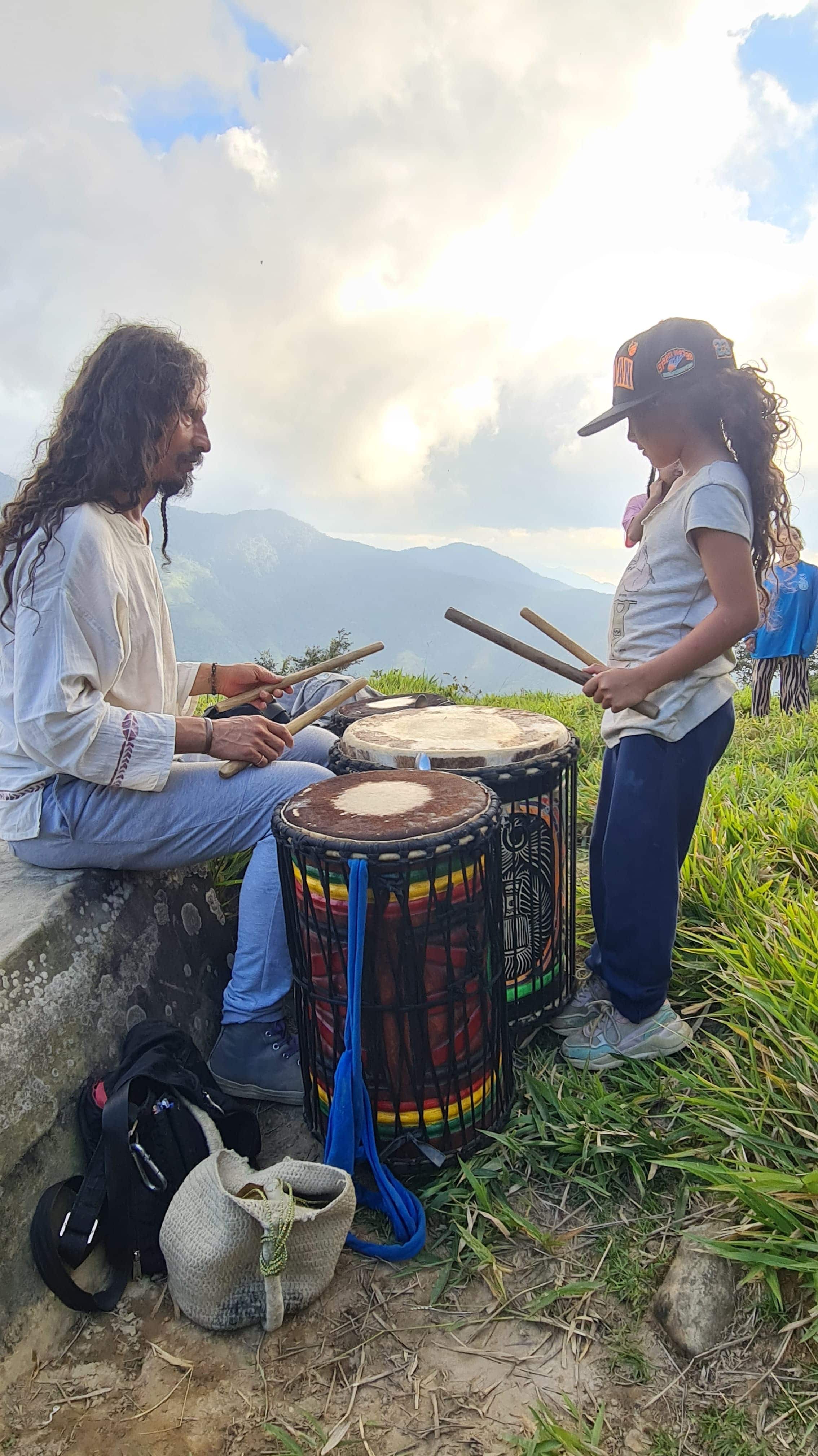 Two people playing drums outdoors on a hillside at sunset.