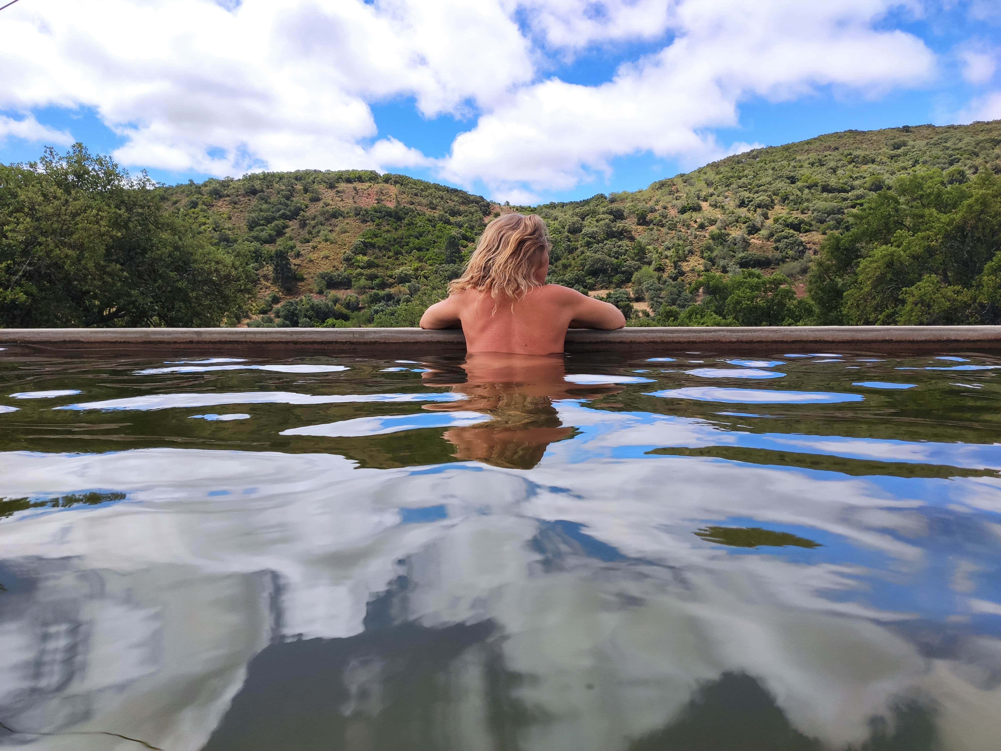 Person in an outdoor natural pool relaxing with blue sky and hills.
