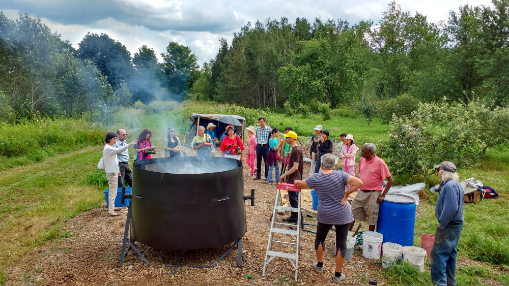 Groups of people gathered around a large outdoor cauldron cooking under trees.