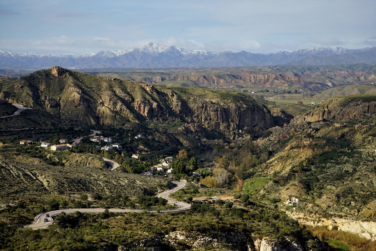 Scenic canyon and winding road through arid hills with snow-capped mountains in the distance.
