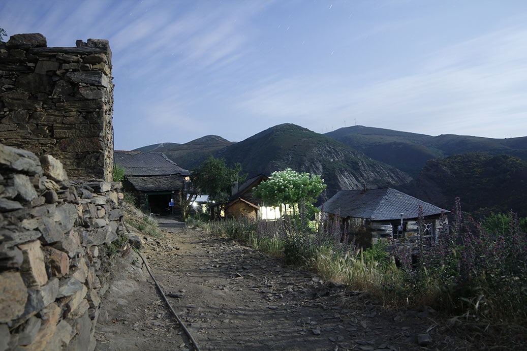 Path through rustic village with stone walls and hills in the background.