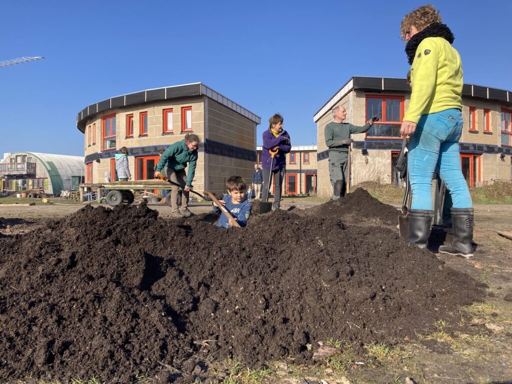 Children and adults building soil mounds on a sunny day at Boekel Ecovillage, with eco-buildings in/.