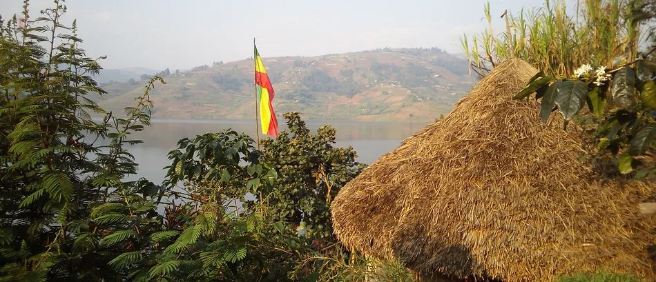Scenic view of a straw hut by Lake Bunyonyi with lush greenery and a flag.