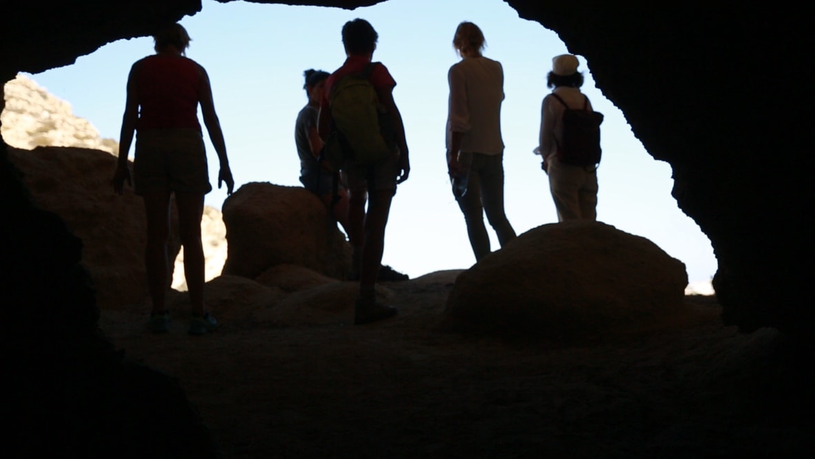 Group of hikers standing at cave entrance with sunlight behind.