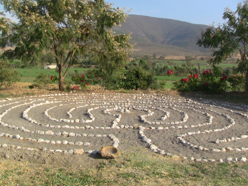 A stone labyrinth laid out in a circular pattern in a rural garden with trees and hills.