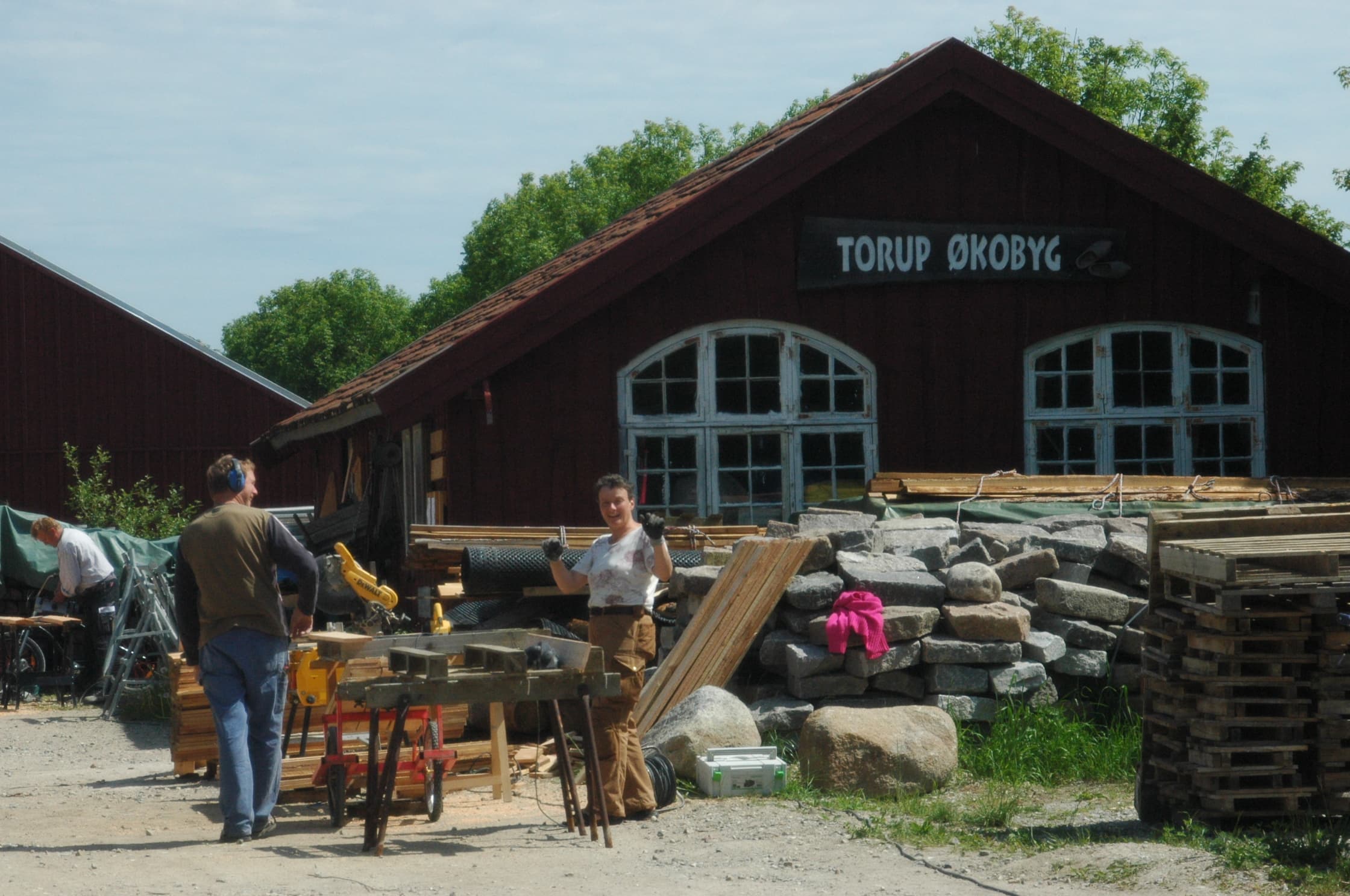 People working with reclaimed wood and tools outside a red wooden building.