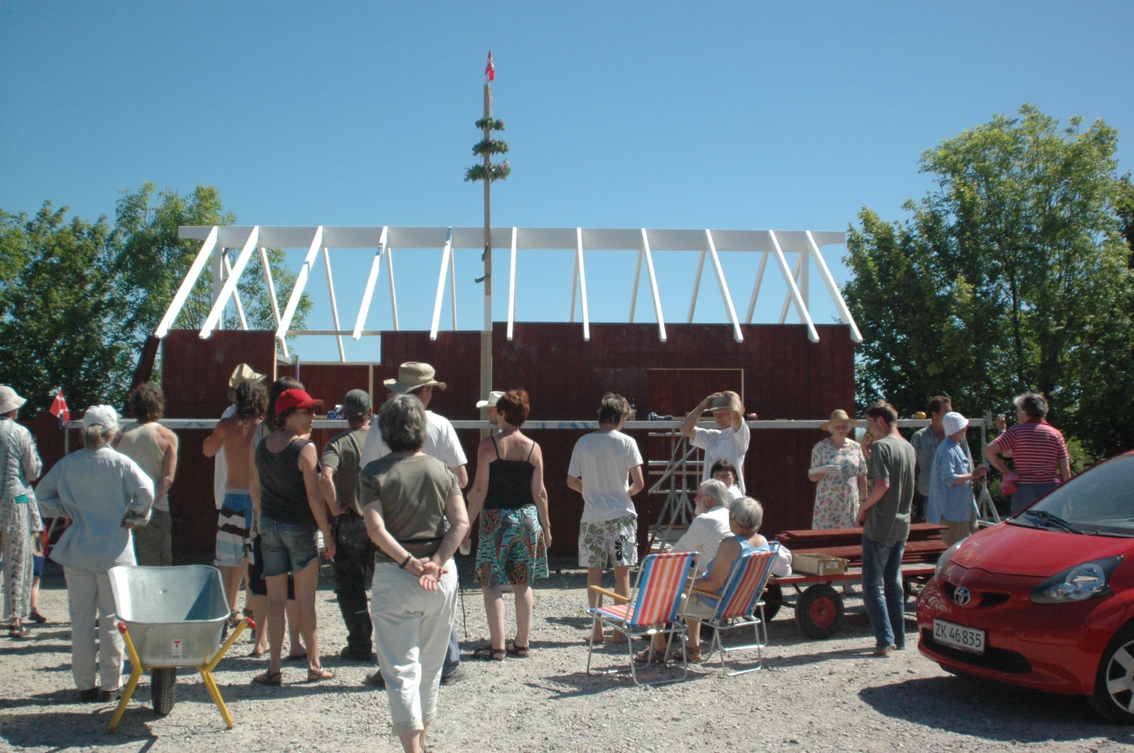People gathered around a building under construction at an eco village site with a red car nearby.