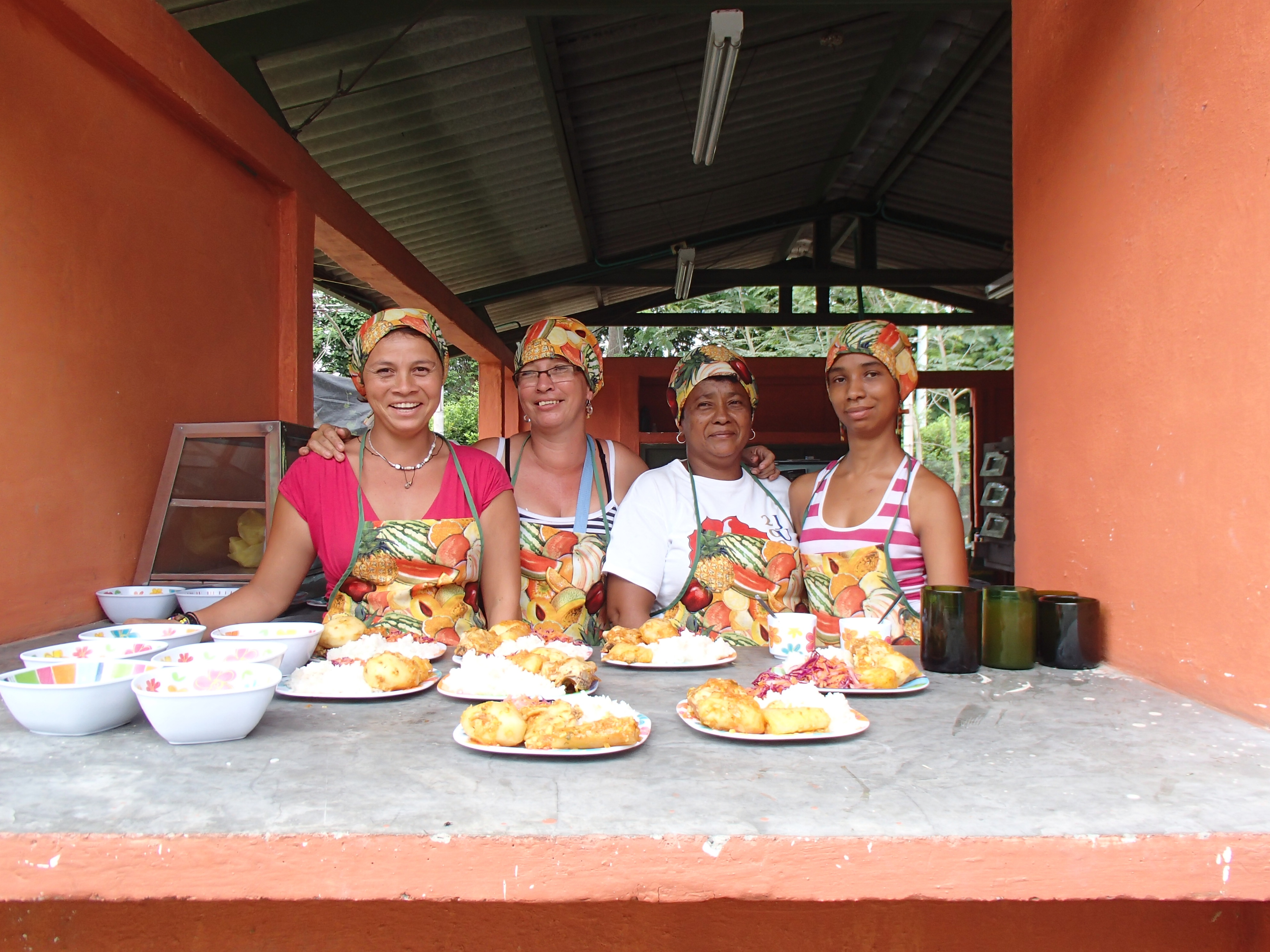 Group of smiling women serving plates of food in an outdoor kitchen.