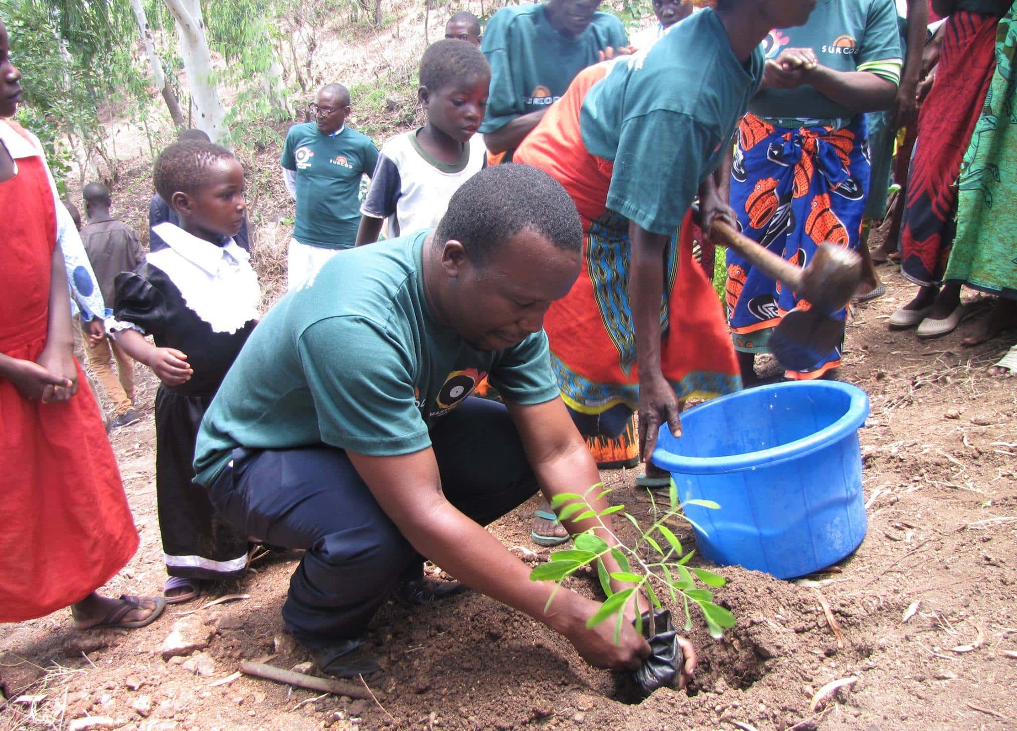Man planting a sapling with community and children around in rural setting.