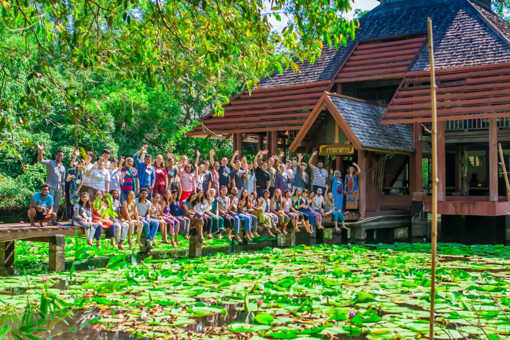 GEN gathering group photo by a pond surrounded by green lily pads and wooden huts.