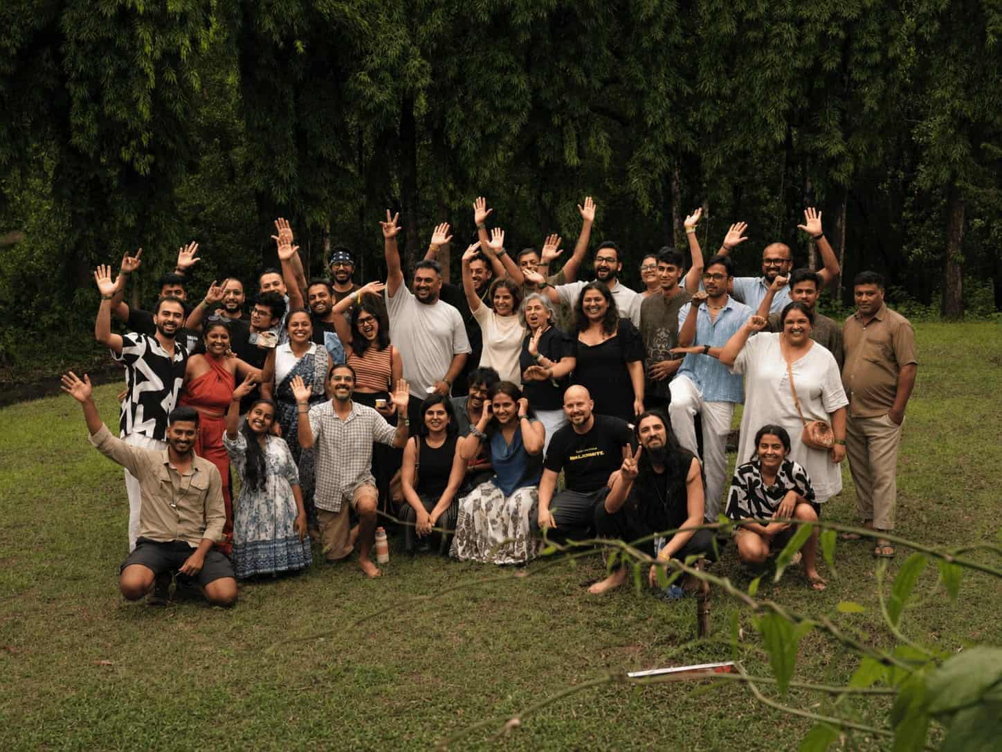Group photo of diverse people outdoors waving at camera in a grassy field.