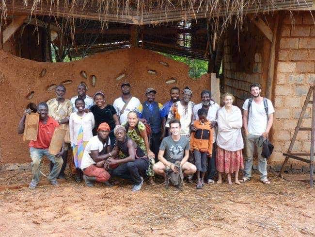 Group of diverse people standing together in front of a thatched building and earthen walls.