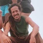 Man with dreadlocks smiling, seated in casual outdoor setting.