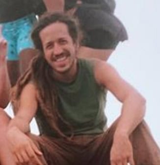 Man with dreadlocks smiling, seated in casual outdoor setting.