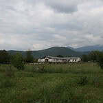 Rural ecovillage farmstead with a long white building amidst greenery under cloudy sky.