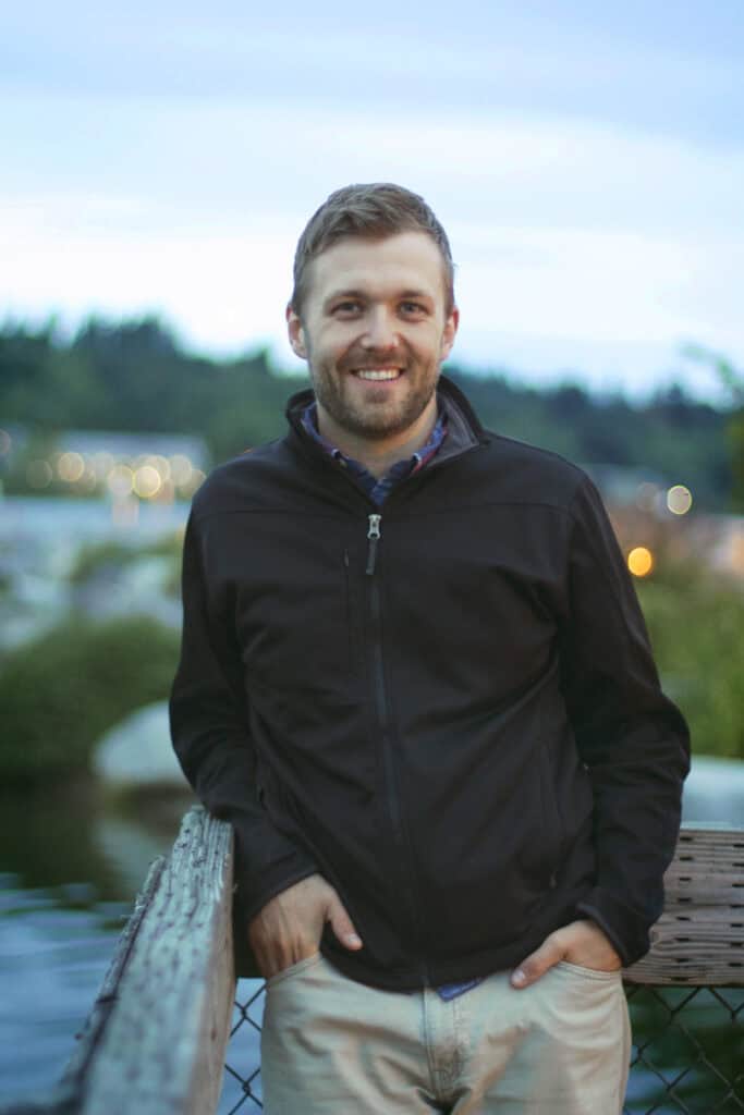 Open air portrait of a smiling man in a black jacket by the water.