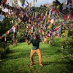 Person in green shirt and hat standing under colorful prayer flags in a grassy field.
