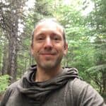 Man smiling outdoors in a forest setting.
