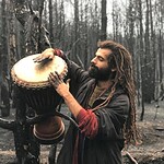 Man with dreadlocks playing a djembe among burnt trees in a forest.