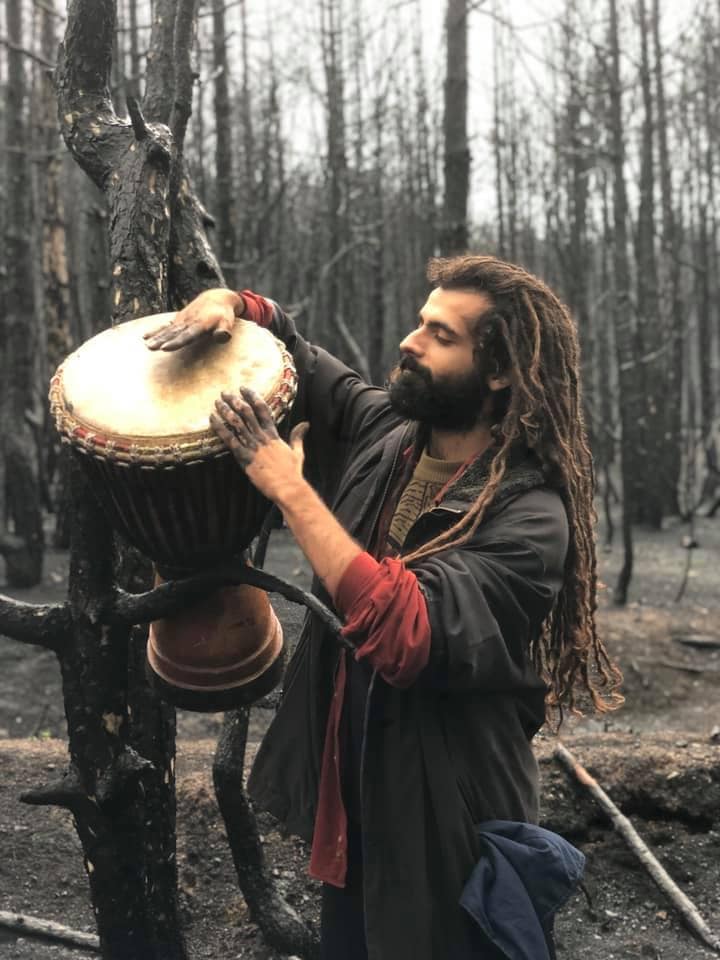 Man with dreadlocks playing a djembe among burnt trees in a forest.