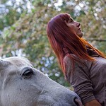 Person with brown hair leaning towards a white horse in an outdoor setting.
