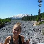 Smiling hiker in a rocky valley with conifer trees and a snow-capped mountain in the distance.