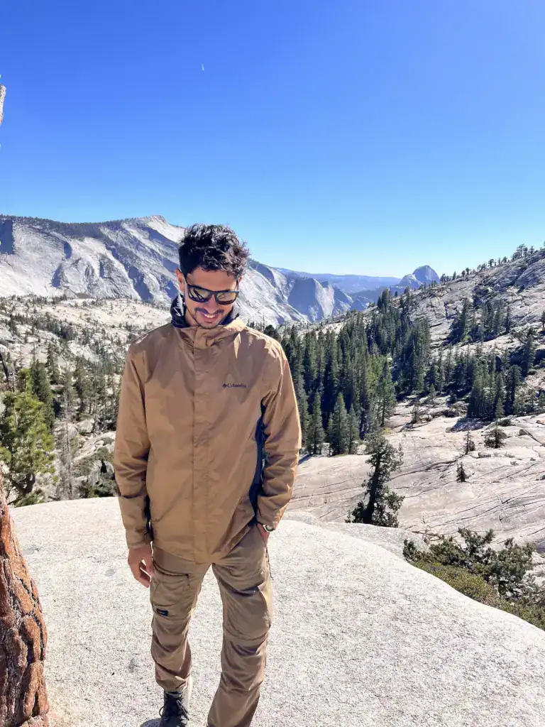 Man in tan outdoor jacket standing on rocky overlook with forest and mountains.