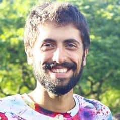 Smiling man in a colorful shirt outdoors with greenery in the background.