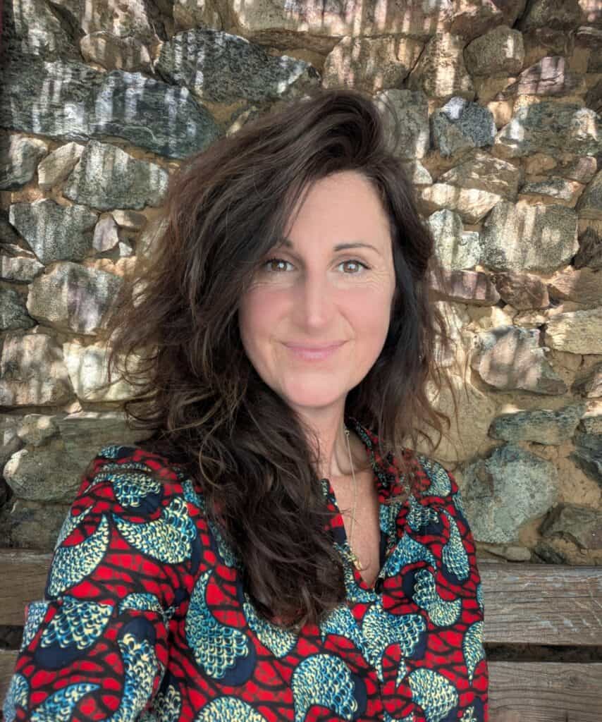 Portrait of a woman with long dark hair, wearing a red patterned blouse, in front of a stone wall.