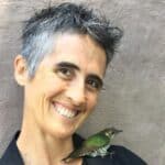 Smiling woman holding a small green bird, outdoors.