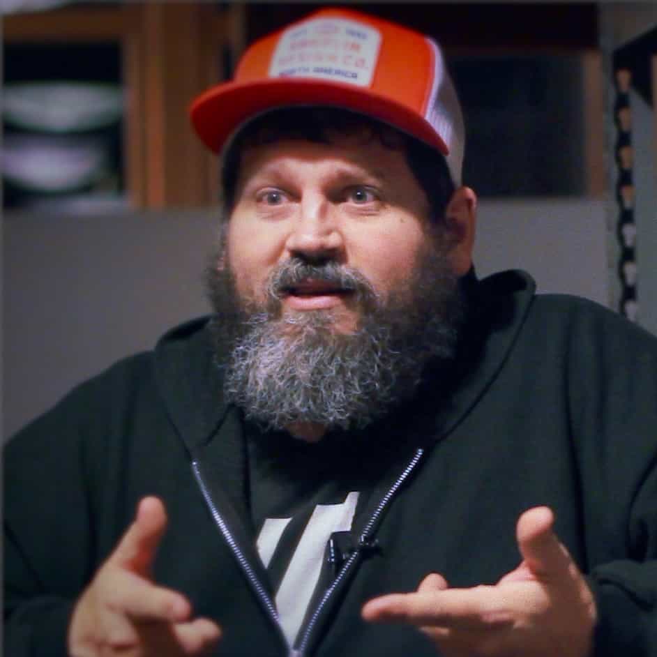 A man with a full beard and wearing a red and white trucker hat is talking animatedly with his hands in an indoor setting, embodying the spirit of Generation Freedom.