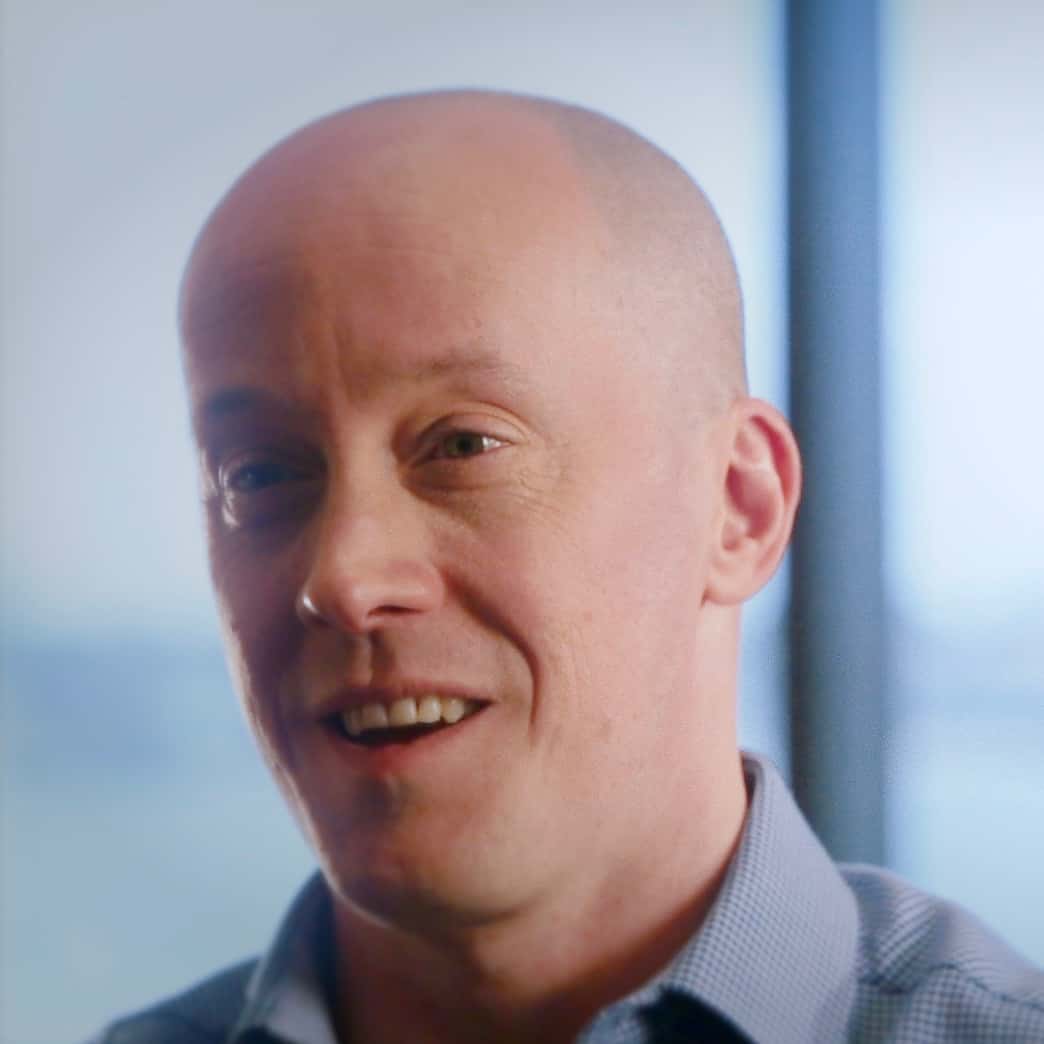 A portrait of a smiling bald man from Generation Freedom with fair skin, wearing a checkered shirt, standing indoors with a soft focus background.