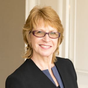 A professional headshot of a smiling woman with shoulder-length blonde hair, wearing glasses, gold earrings, and a black blazer. The background is a softly lit, neutral-colored room.