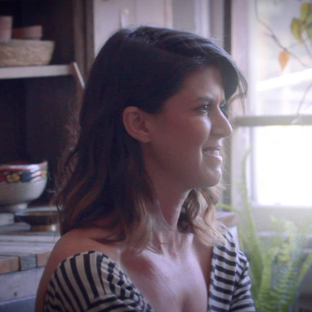A woman with shoulder-length hair, wearing a striped top from Generation Freedom, smiles while looking to the side in a warmly lit room filled with plants.